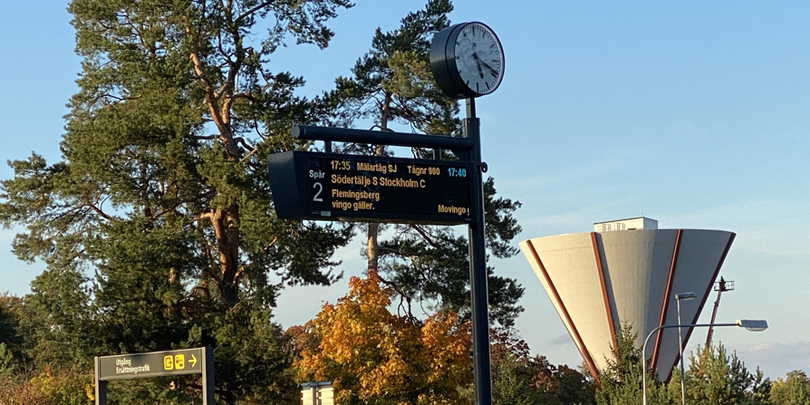 Skylt med tågets avgångstider vid stationen i Nykvarn. I bakgrunden syns vattentornet. 